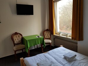 Une chambre d’hôtel avec un lit, une table recouverte d’une nappe verte, deux chaises, un téléviseur fixé au mur et une fenêtre avec des rideaux.