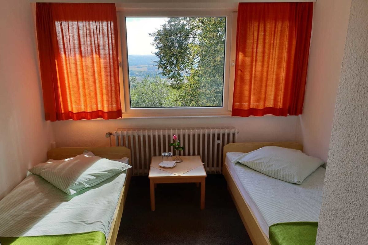 Een kleine kamer met twee enkele bedden, een tafeltje met een bloemenvaas en een raam met gordijnen.