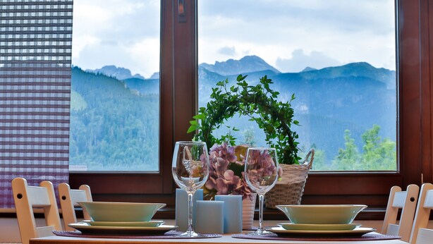 A dining table with two plates, two wine glasses, and candles, set against a window with a mountain view. - VIP Apartamenty Widokowe (Zakopane)