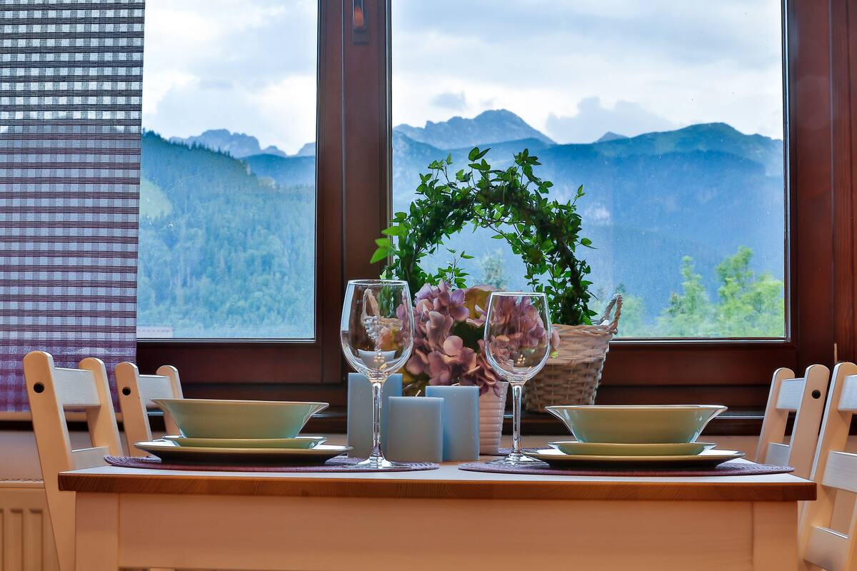 A dining table with two plates, two wine glasses, and candles, set against a window with a mountain view.