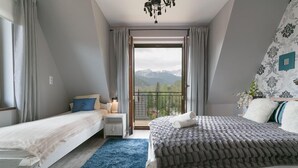 A bedroom with a large bed, a smaller bed, a nightstand, a lamp, a blue rug, and a view of mountains through a balcony. - VIP Apartamenty Widokowe (Zakopane)