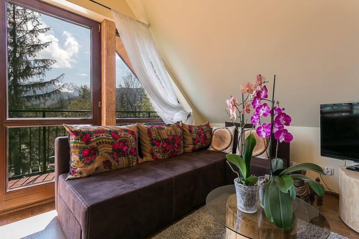 A living room with a brown sofa, colorful pillows, a glass coffee table, and a potted orchid plant.