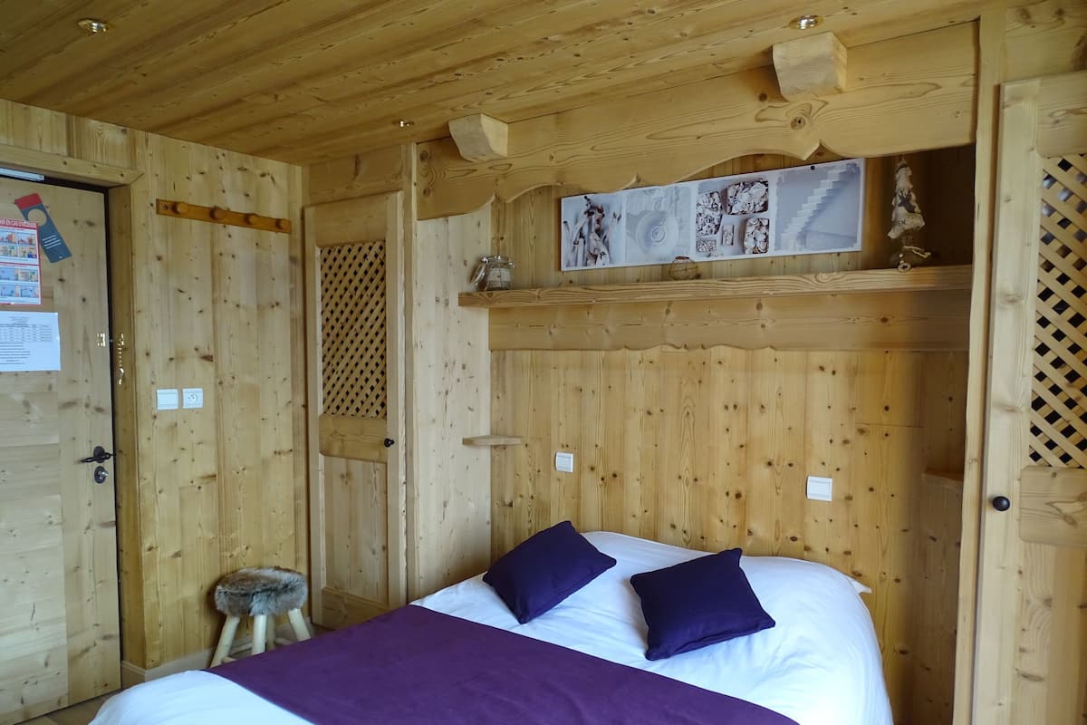 A wooden room with a bed, a stool, and a shelf with decorative items.
