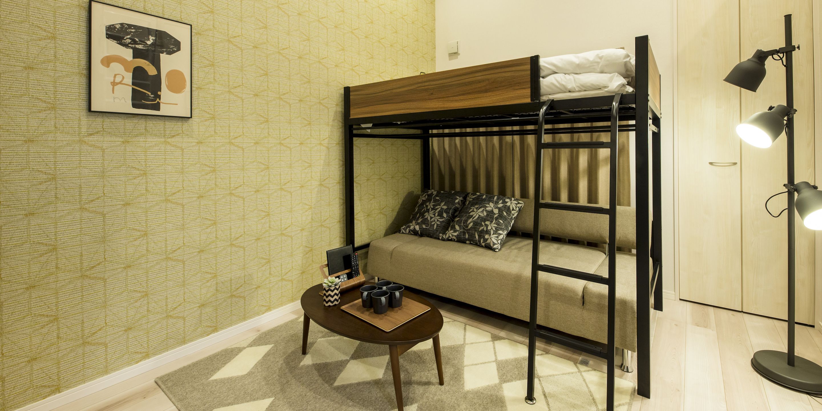 A bunk bed with a wooden headboard, a sofa with patterned cushions, a small table with a plant and a tray of mugs, and a floor lamp.