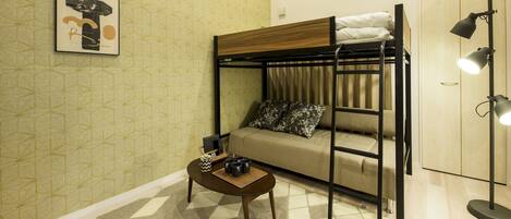 A bunk bed with a wooden headboard, a sofa with patterned cushions, a small table with a plant and a tray of mugs, and a floor lamp.