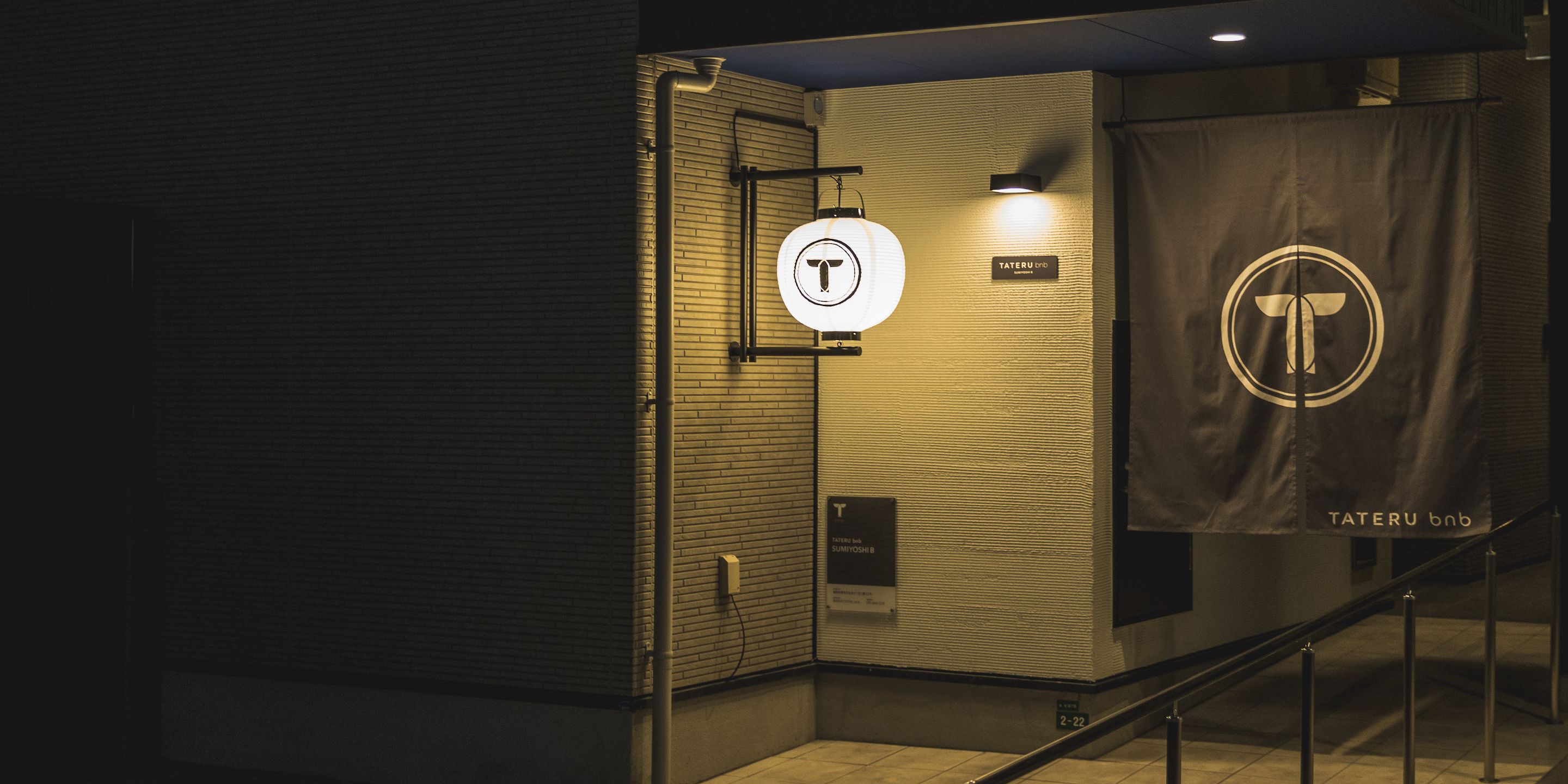 Ingresso di un edificio moderno con un cartello TATERU bnb, un logo a forma di T e una insegna illuminata a parete.