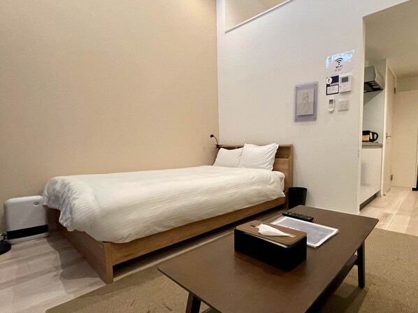 Un lit simple avec un cadre en bois et du linge de lit blanc, une table de chevet avec une télécommande et une boîte de mouchoirs, et un panneau Wi-Fi fixé au mur.