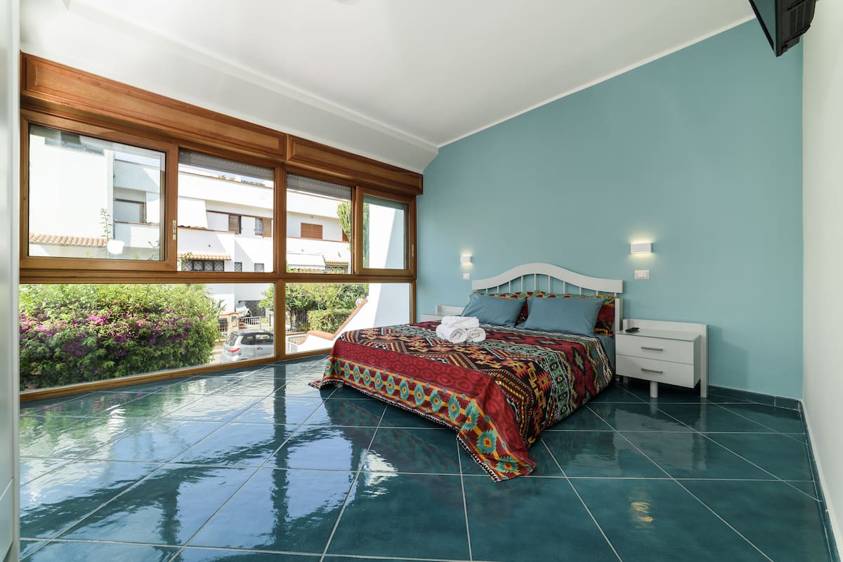 A bedroom with a large window, a bed with a patterned bedspread, a nightstand, and a white drawer unit.