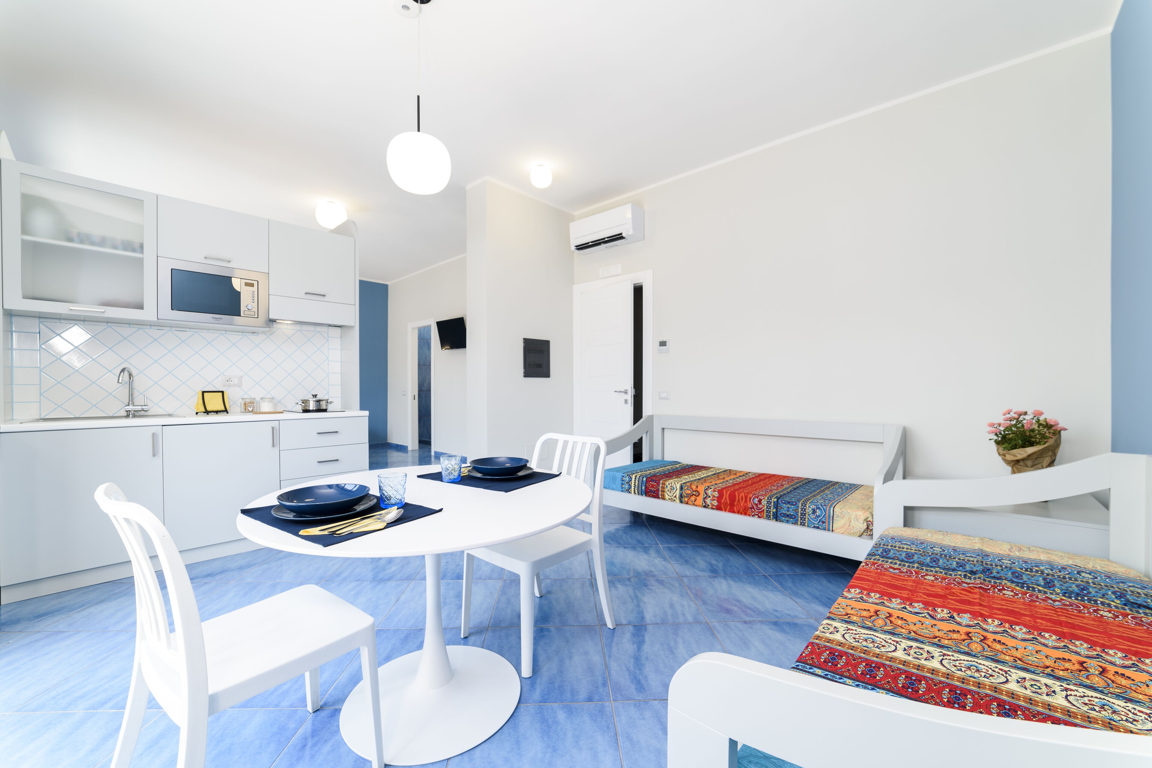 A modern kitchen with white cabinets, a microwave, and a sink. There is a dining table with chairs and a colorful rug.