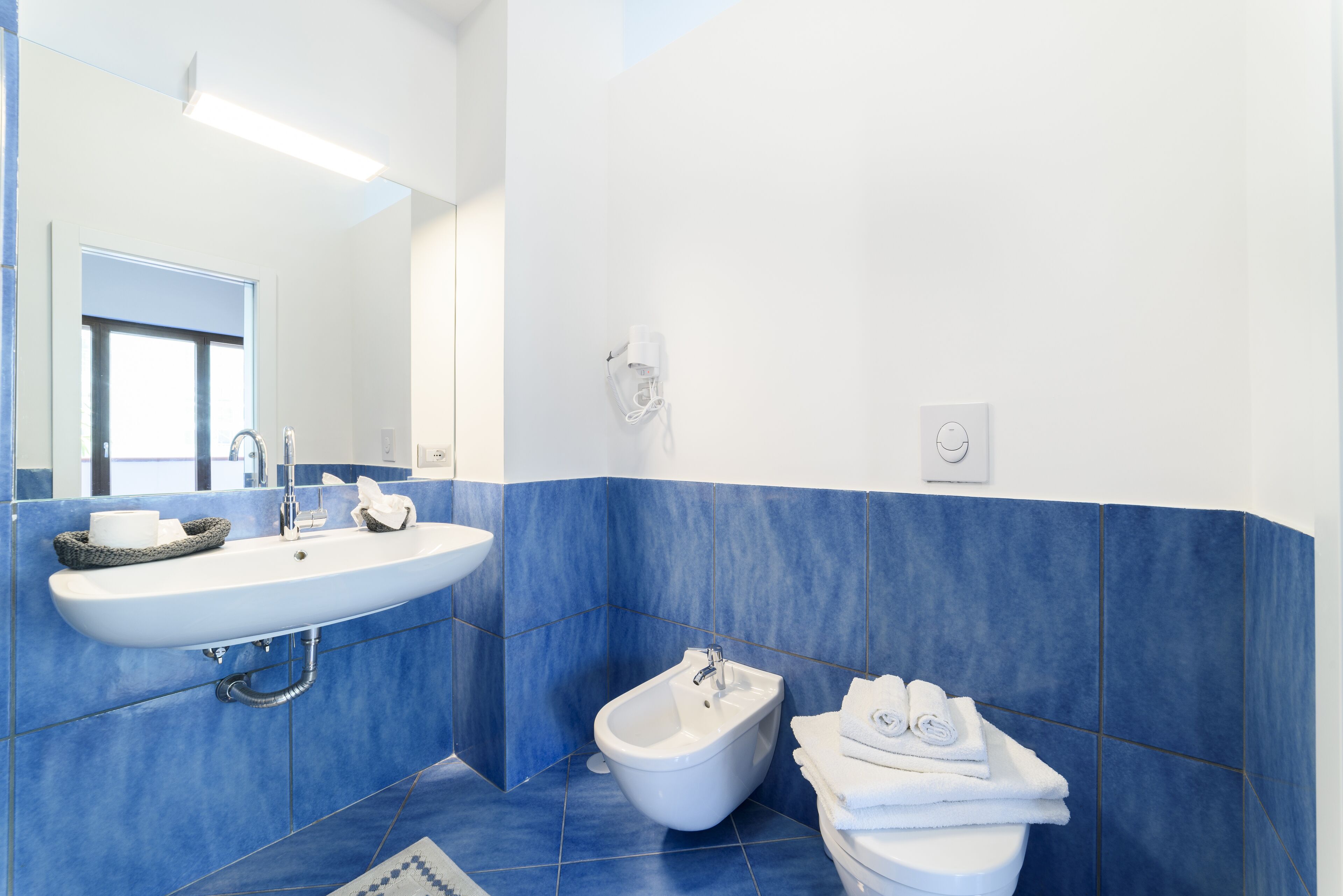 A modern bathroom with a white sink, bidet, and toilet, blue tiles, and a mirror.