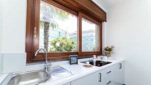 Una cocina moderna con un lavamanos de acero inoxidable, armarios blancos, una ventana con vistas a edificios y árboles, y una pequeña encimera con una cafetera y una planta.
