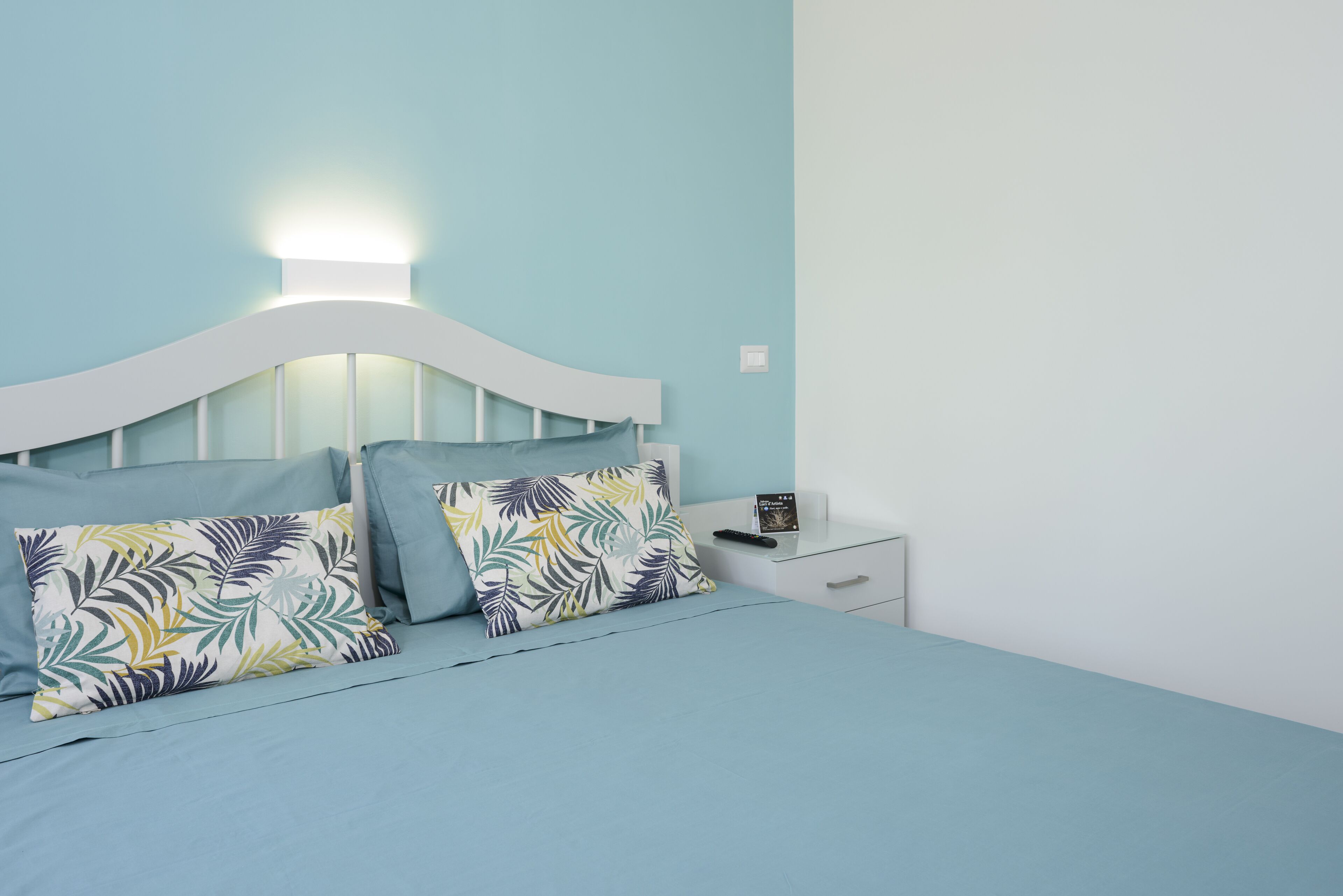 A bed with a white headboard, two pillows with a tropical leaf pattern, and a bedside table with a remote control.