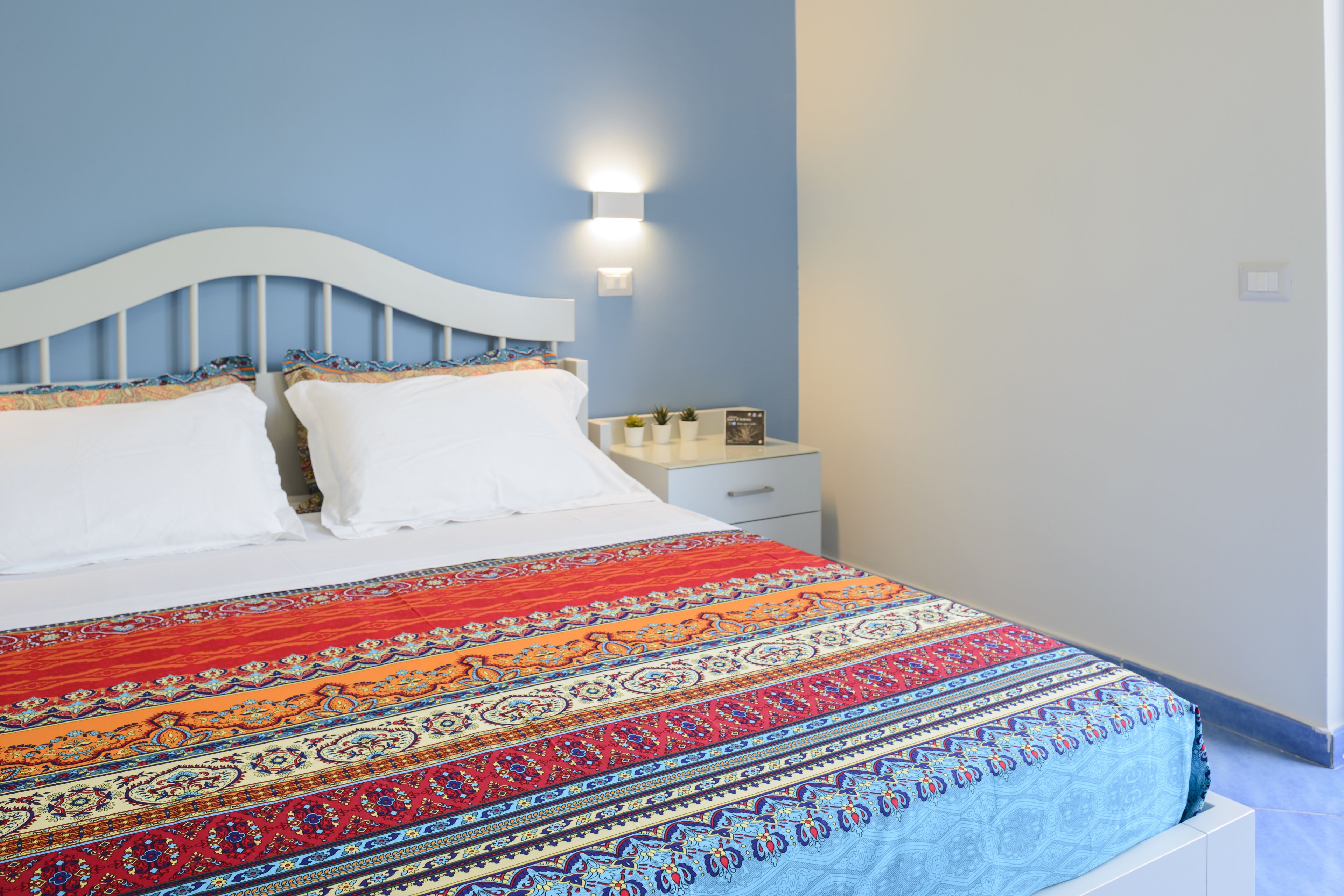 A bed with a patterned bedspread, a nightstand with a lamp, and a wall-mounted light fixture.