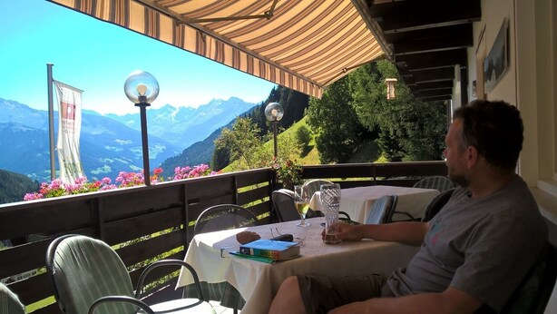 A man sitting at a table with a mountain view, a flag, and a lamp post. - Jägerhof Geniesser & Wanderhotel (San Leonardo in Passiria)