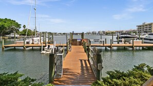 A wooden pier leading to a marina with boats docked, surrounded by greenery and buildings.