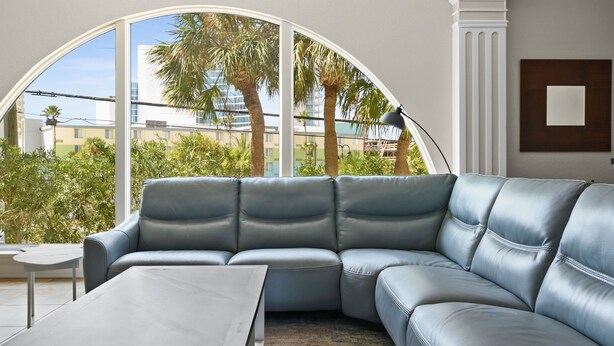 A modern living room with a large L-shaped sectional sofa, a small round table, and a view of palm trees and buildings through a large arched window.
