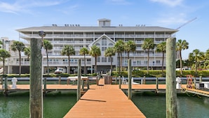 A wooden pier leading to a multi-story hotel with balconies and palm trees.