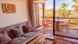 A modern living room with a sofa, a coffee table, and a view of a balcony with outdoor seating and palm trees. - Enjoy the Ocean View and take a stroll on the Stunning Private Beach (Cabo San Lucas)