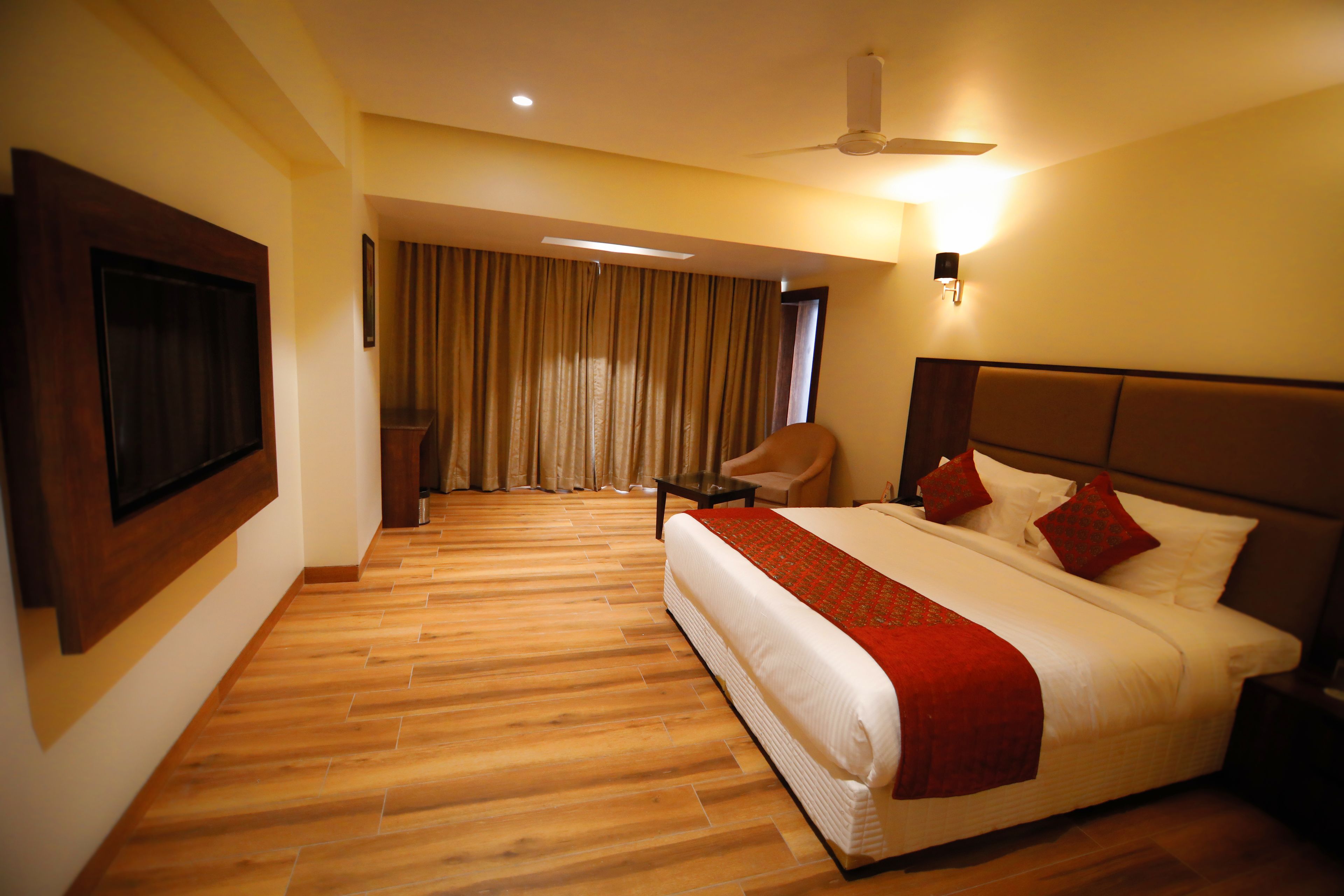 A hotel room with a large bed, a flat-screen TV mounted on the wall, a ceiling fan, and a seating area with a chair and curtains.
