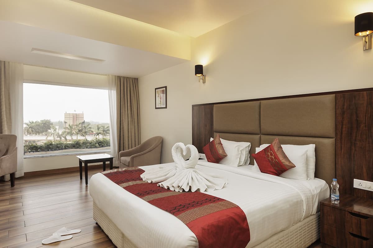 Una habitación de hotel con una cama grande, vistas a palmeras y un edificio, una silla, una mesa y una mesita de noche con una botella.