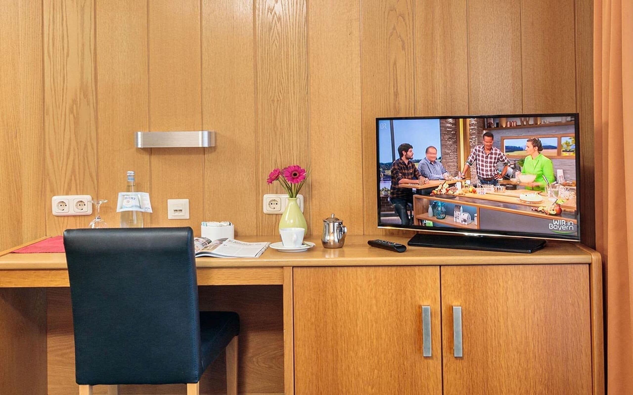 Un escritorio de madera con un televisor que muestra un programa de cocina, un florero con flores, un control remoto y una silla.