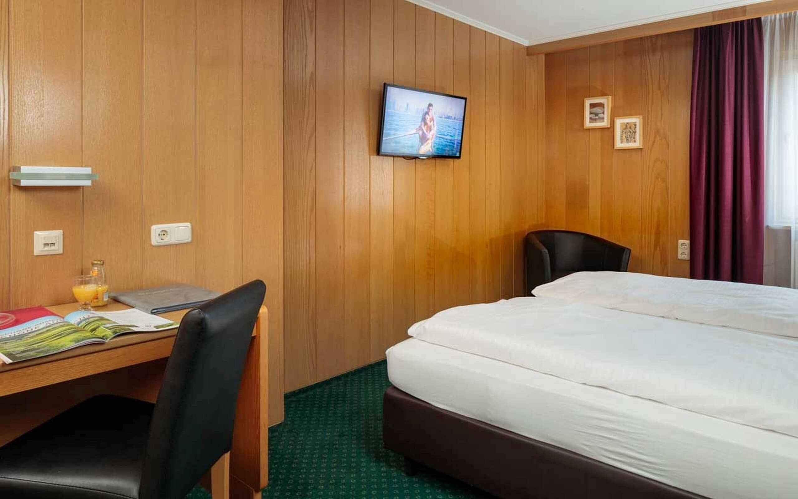 A hotel room with a wooden paneled wall, a television mounted above a desk, a bed with white bedding, and a chair.