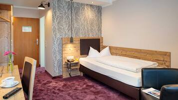 A hotel room with a wooden headboard, a bed with white linens, a desk with a chair, a small table with a phone and a cup of coffee, and a wall with a tree pattern.