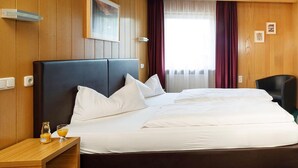 A hotel room with a large bed, wooden panel walls, a window with curtains, a small table with a bottle and glass, and a chair. - Hotel Gasthof Pockinger Hof (Pocking)