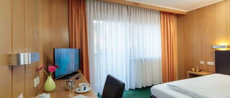 Habitación de hotel con pared de paneles de madera, una cama, un escritorio con televisor, un florero con flores, una taza de café y un periódico.