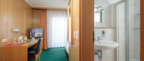 A hotel room with a wooden desk, a television, a green carpet, a shower, and a white sink.