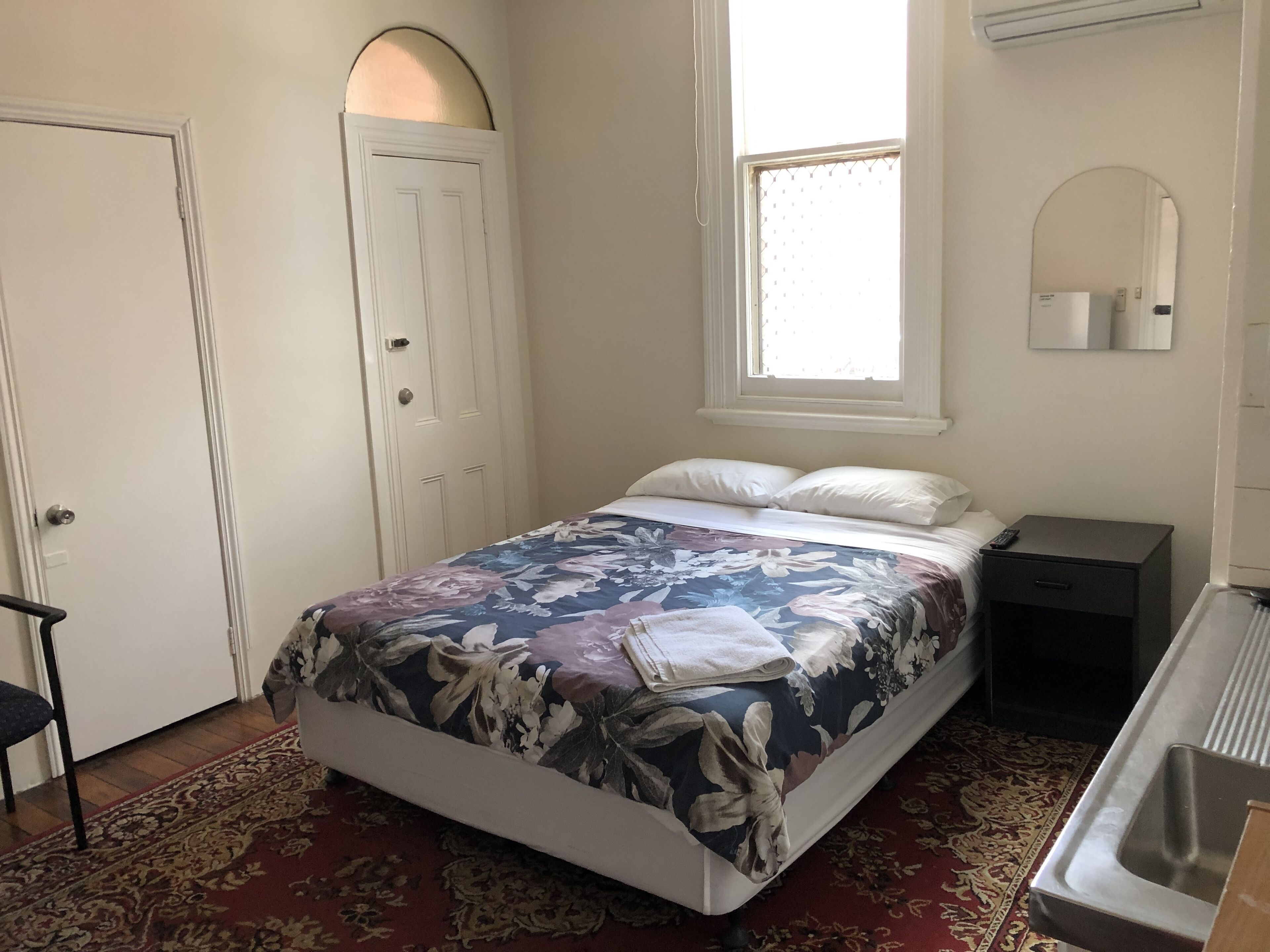 Un dormitorio con cama, mesita de noche, ventana, puerta y espejo.