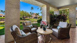 A covered outdoor seating area with wicker furniture and a round table, overlooking a landscaped garden with a pond and palm trees. - 🏖️Ground Floor Condo | Lagoon Views + Large Lanai + A/C (Waikoloa)