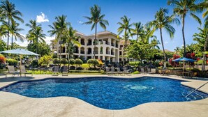 A swimming pool with lounge chairs and umbrellas, surrounded by palm trees and a building with multiple balconies. - 🏖️Ground Floor Condo | Lagoon Views + Large Lanai + A/C (Waikoloa)