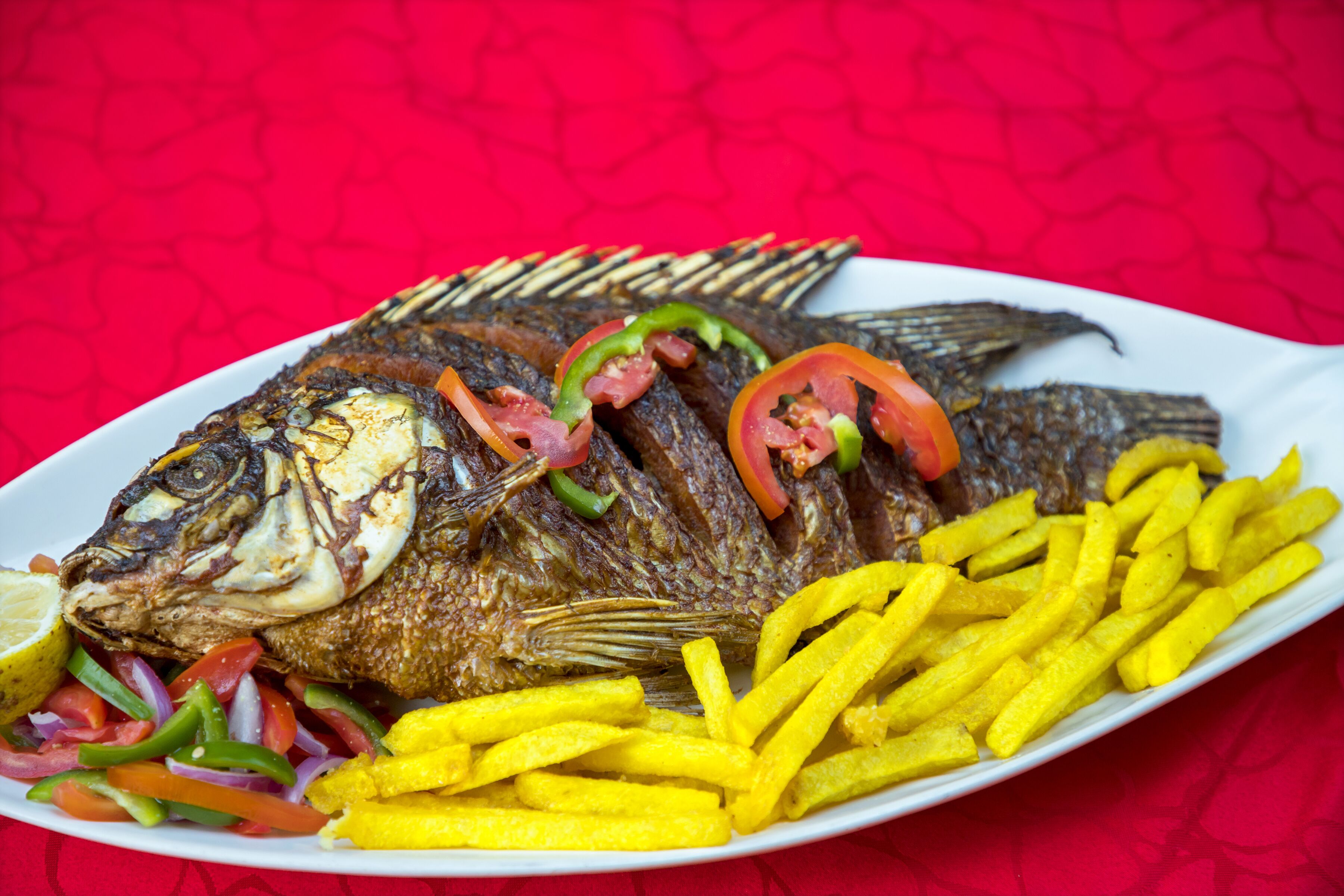 Un poisson entier grillé, garni de lamelles de poivrons et d’oignons, servi avec des frites dans une assiette blanche.