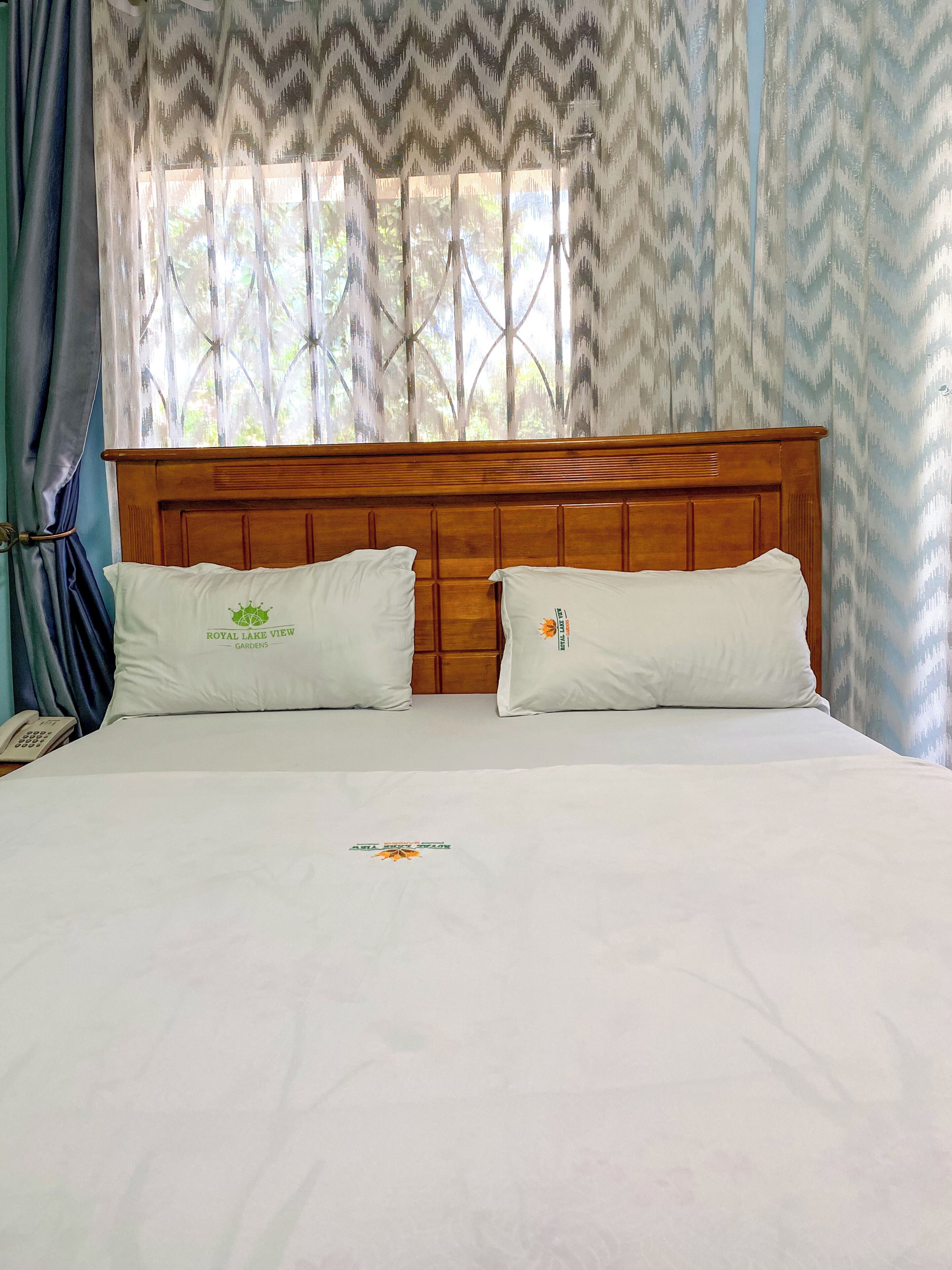 Una cama con dos almohadas, un cabecero de madera y una ventana con cortinas estampadas.