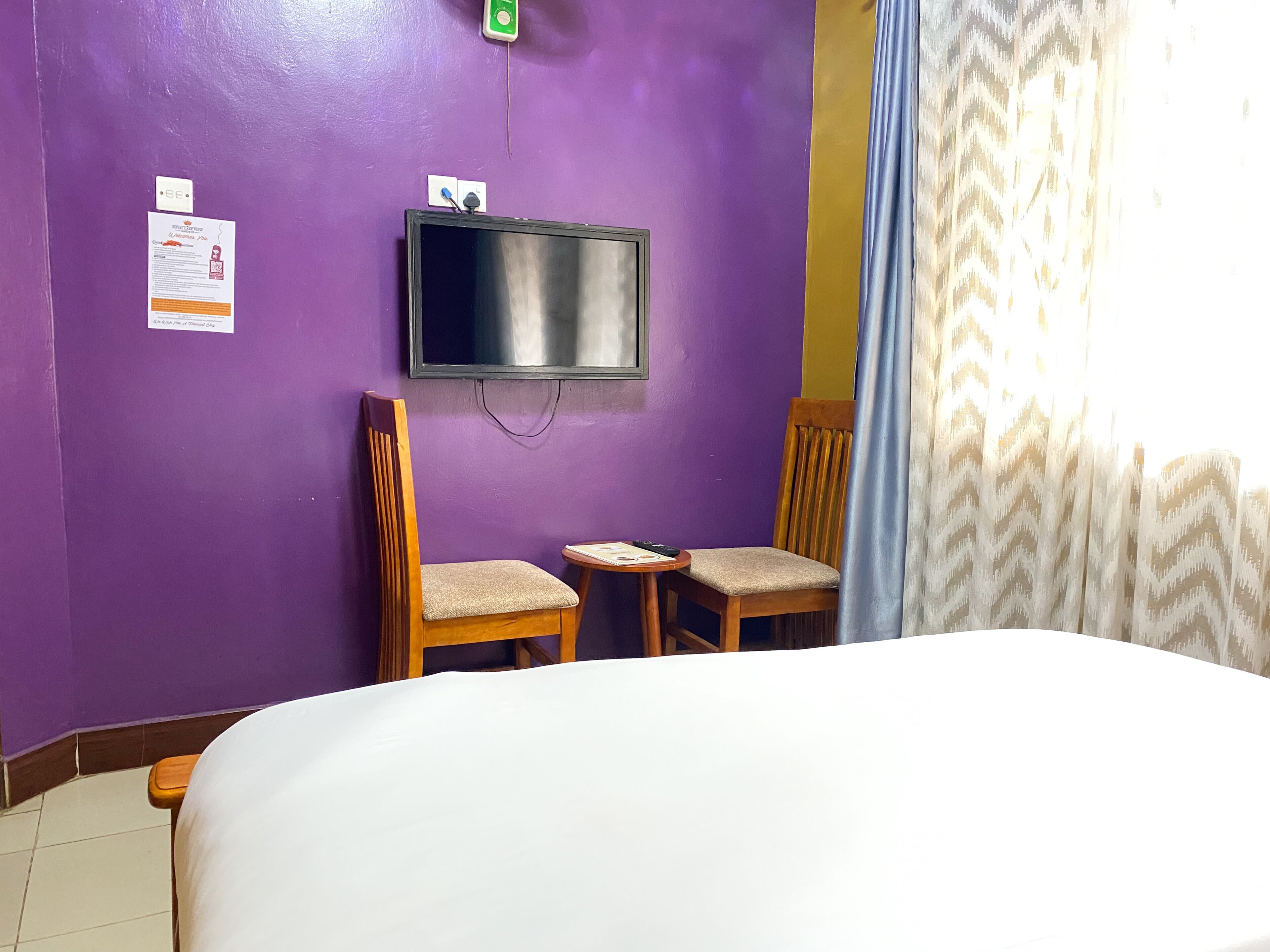 Une pièce comprenant un lit, deux chaises en bois, une petite table, un téléviseur fixé au mur et un rideau.
