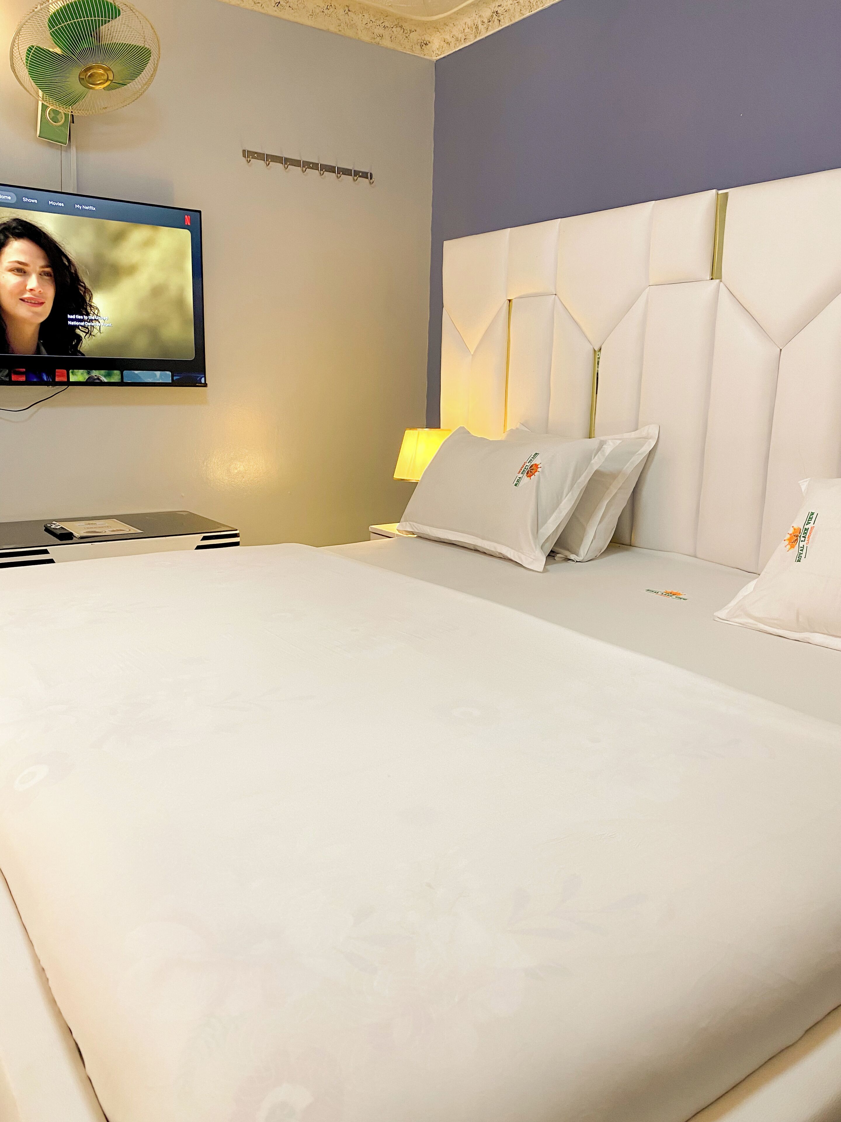 A hotel room with a bed, a TV mounted on the wall, a desk with a lamp, and a ceiling fan.