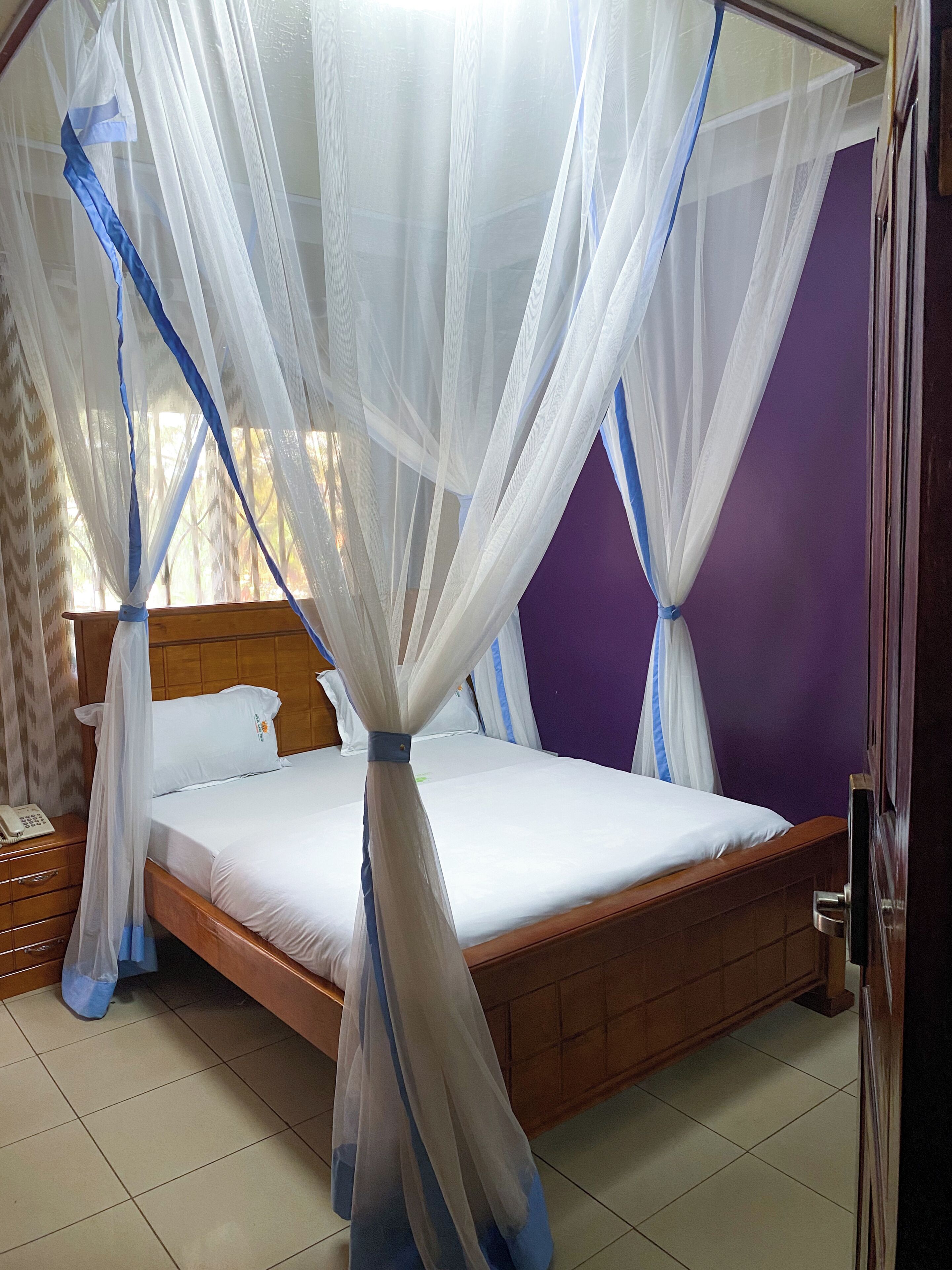 A bed with a canopy, a wooden headboard, a bedside table with a phone, and a purple wall.