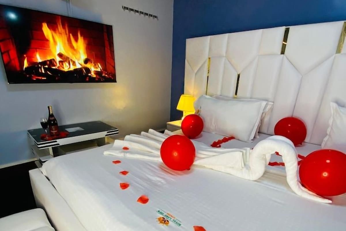 A hotel room with a large bed, a flat-screen TV mounted on the wall, a bedside table with a bottle and glasses, and a wall-mounted fan.