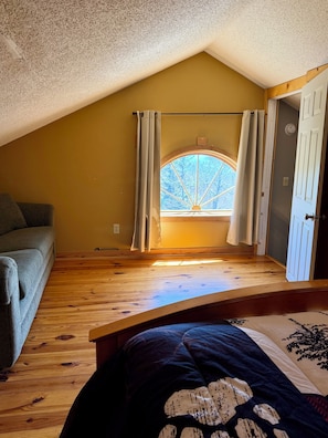 Une chambre située dans un espace de type « loft », avec un plafond en pente, une fenêtre avec des rideaux, un plancher en bois et un lit recouvert d’une couverture à motifs.