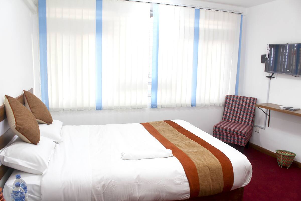 Habitación de hotel con una cama grande, una silla, un televisor y una ventana con persianas.