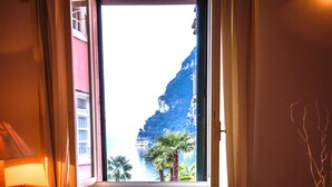A room with a large window overlooking a coastal area, a radiator, a wicker chair, and a striped bed. - Queen of the Lake (Riva del Garda)