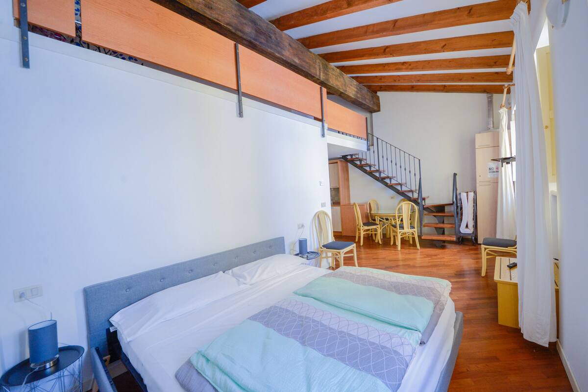 A bedroom with a bed, a desk, and a chair. There is a staircase leading to an upper level.