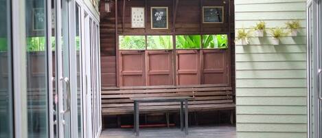 Un espacio de estar al aire libre con techo, con bancos de madera y una mesa, rodeado por puertas de vidrio y una pared verde.