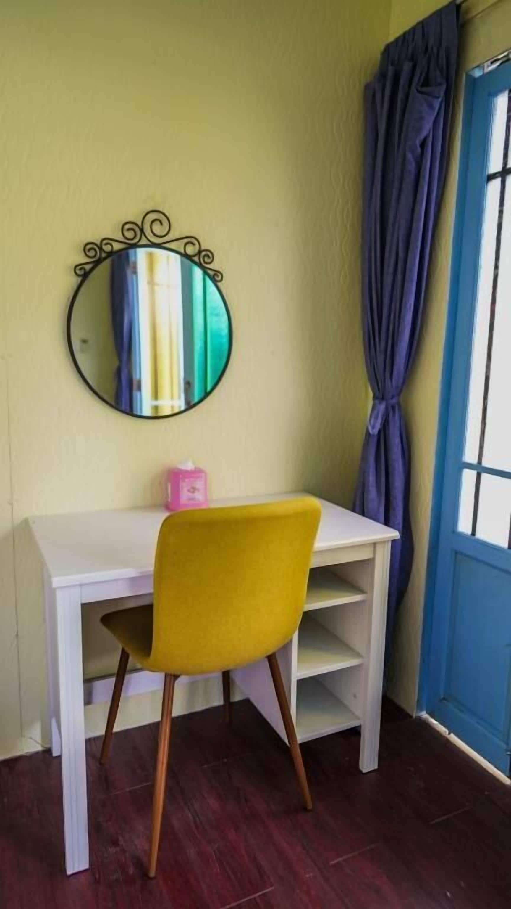 A room with a white desk, a yellow chair, a round mirror with a black frame, and a blue door.