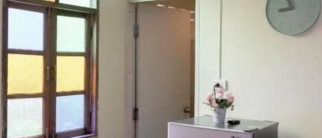A small, empty room with a single window, a wall clock, a refrigerator, and a trash bin.