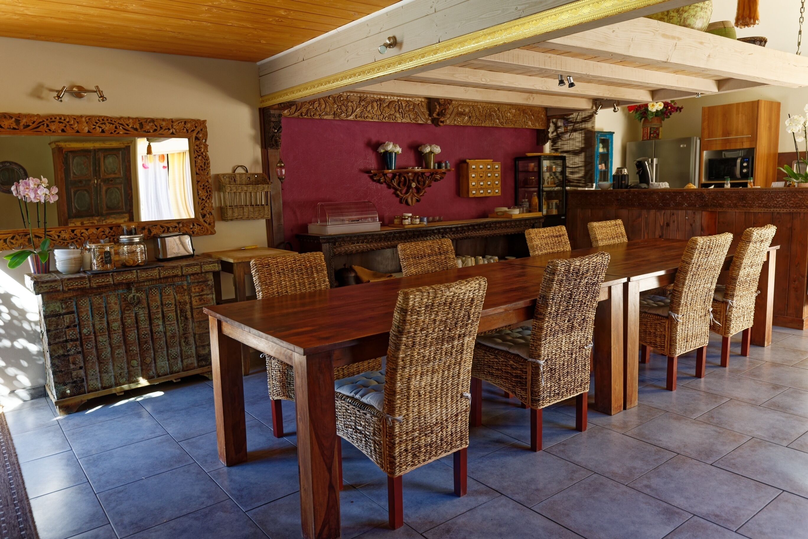 A dining area with a long wooden table and wicker chairs, a kitchen in the background, and a decorative mirror above the table.