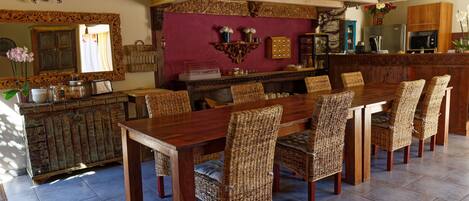 Un comedor con una mesa de madera larga y sillas de mimbre, una cocina al fondo y un espejo decorativo sobre la mesa.