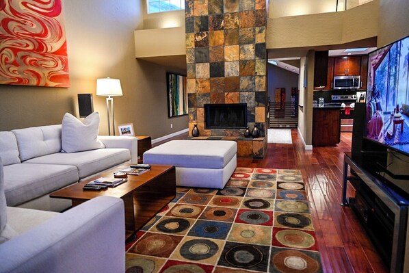 Una sala de estar moderna con sofá, reposapiés, mesa de centro y televisor. Incluye una chimenea, una cocina al fondo y una alfombra estampada.
