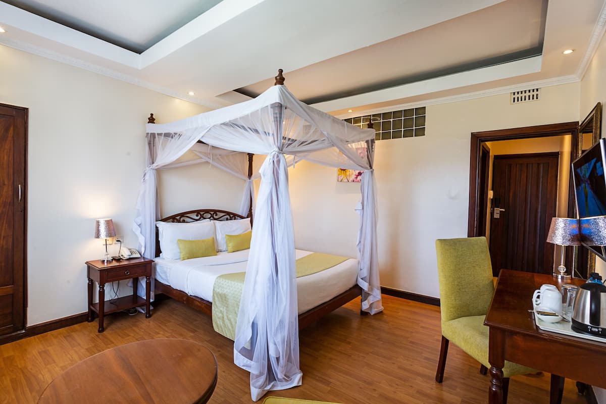 A bedroom with a four-poster bed, a bedside table with a lamp, a green armchair, and a wooden coffee table.